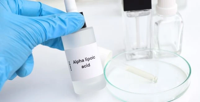 Flesje met etiket “Alpha lipoic acid” (alfa-liponzuur) in blauwe latexhandschoen, naast petrischaal en druppelpipet in laboratoriumsetting.